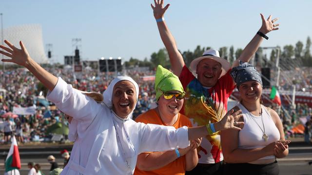 Eine Gruppe von jungen Menschen reißt die Arme in die Höhe. Sie lachen. Hinter ihnen sieht man auf einem Frei-Gelände eine Menschen-Menge.