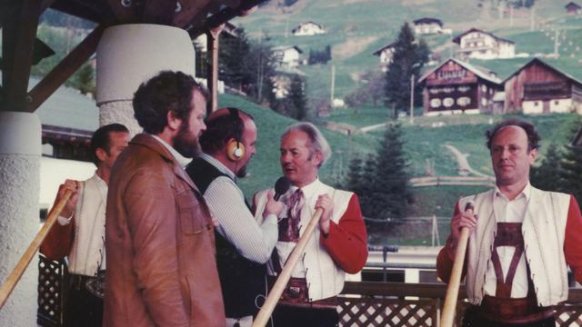 Zwei Reporter interviewen drei Alphornbläser in traditioneller Kleidung vor alpiner Kulisse