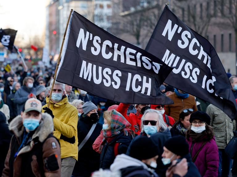 Menschen demonstrieren gegen Coronamaßnahmen und tragen schwarze Fahnen mit der Aufschrift „N’SCHEISS MUSS ICH“.