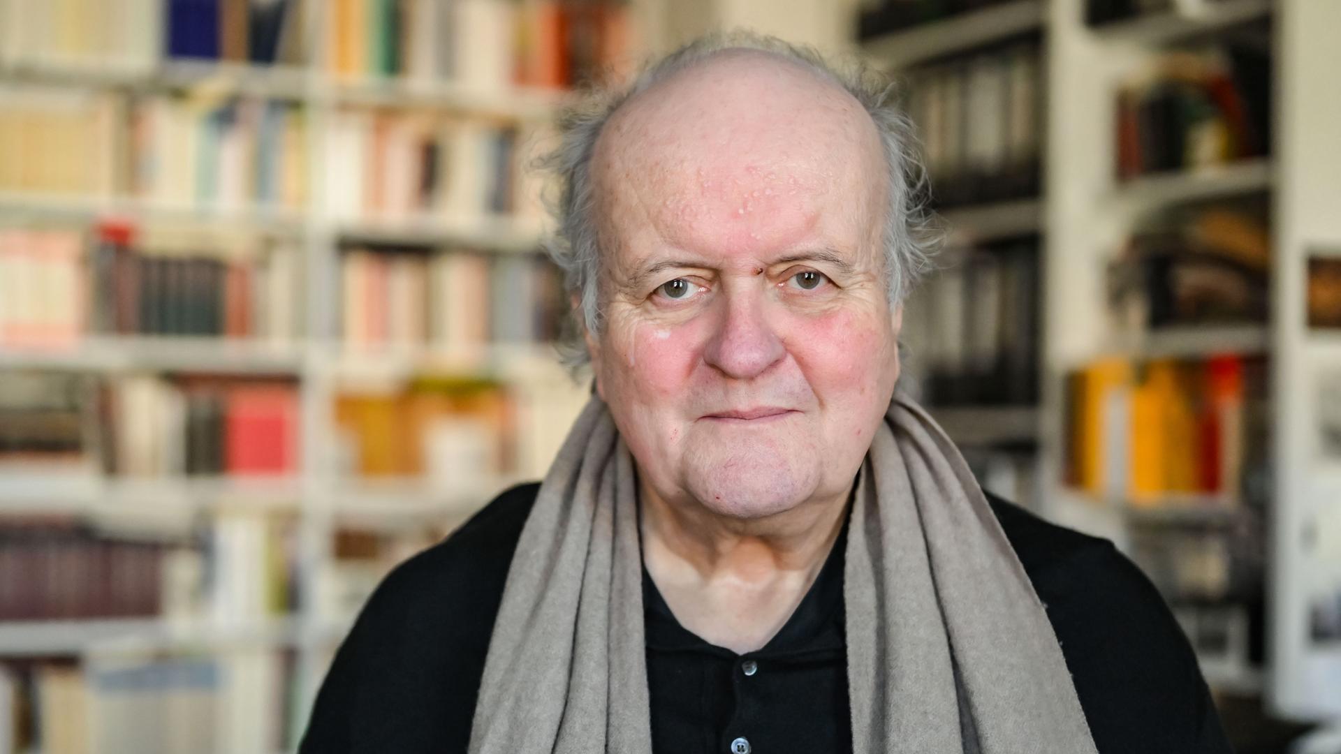 Wolfgang Rihm, ein älterer Mann, blickt freundlich in die Kamera. Er trägt ein schwarzes Hemd und einen braunen Schal. Hinter ihm ist ein großes Bücherregal zu sehen.