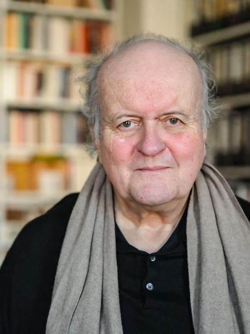Wolfgang Rihm, ein älterer Mann, blickt freundlich in die Kamera. Er trägt ein schwarzes Hemd und einen braunen Schal. Hinter ihm ist ein großes Bücherregal zu sehen.