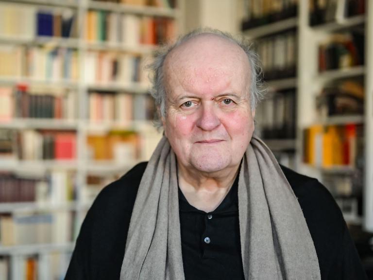 Wolfgang Rihm, ein älterer Mann, blickt freundlich in die Kamera. Er trägt ein schwarzes Hemd und einen braunen Schal. Hinter ihm ist ein großes Bücherregal zu sehen.