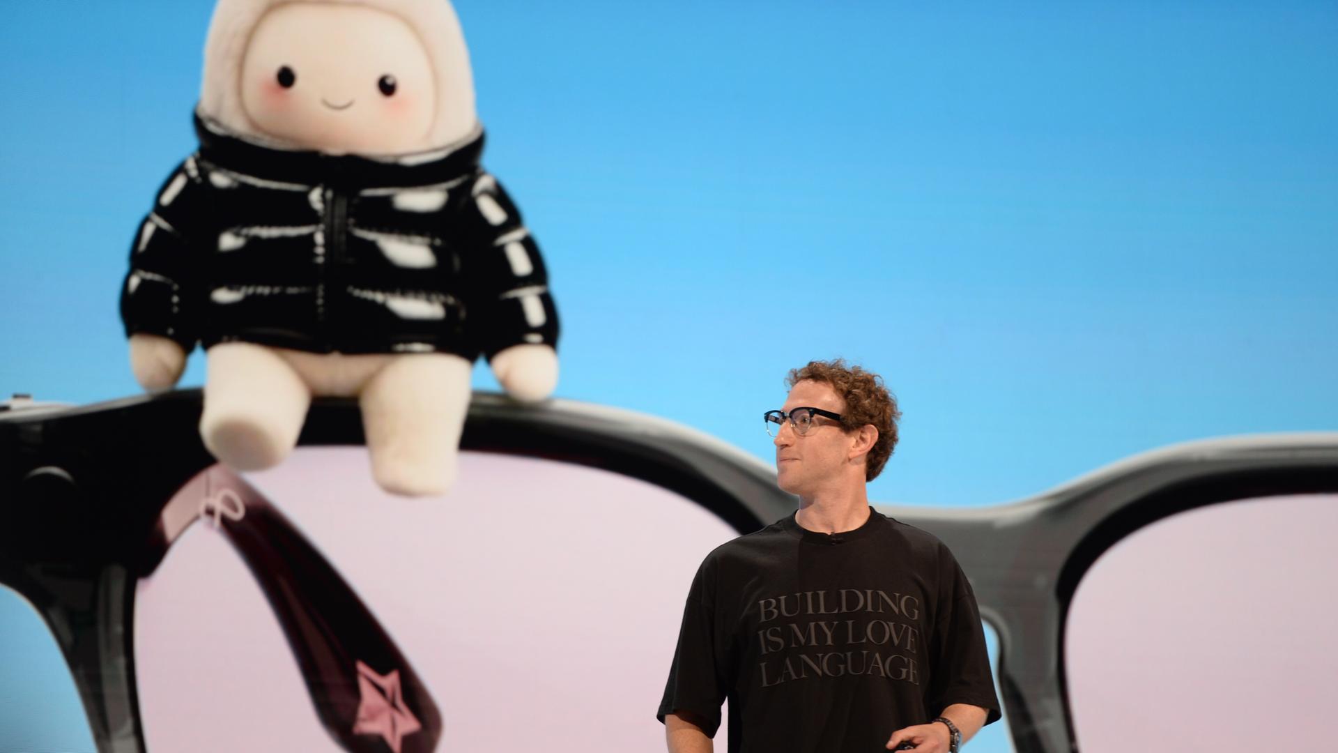 Mark Zuckerberg, Chef des Facebook-Konzerns Meta, steht auf einer Bühne und trägt ein schwarzes T-Shirt mit einer Aufschrift. Hinter ihm ist auf einem Bildschirm ein riesiges Wesen zu sehen, das an ein Tamagotchi oder ein Labubu erinnert.