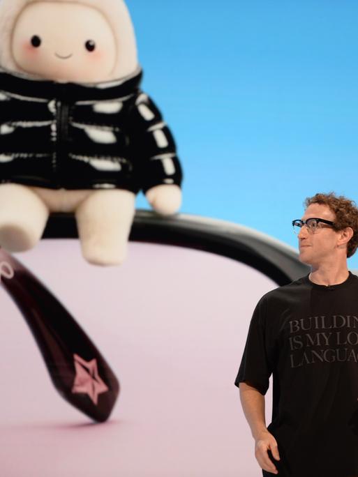 Mark Zuckerberg, Chef des Facebook-Konzerns Meta, steht auf einer Bühne und trägt ein schwarzes T-Shirt mit einer Aufschrift. Hinter ihm ist auf einem Bildschirm ein riesiges Wesen zu sehen, das an ein Tamagotchi oder ein Labubu erinnert.