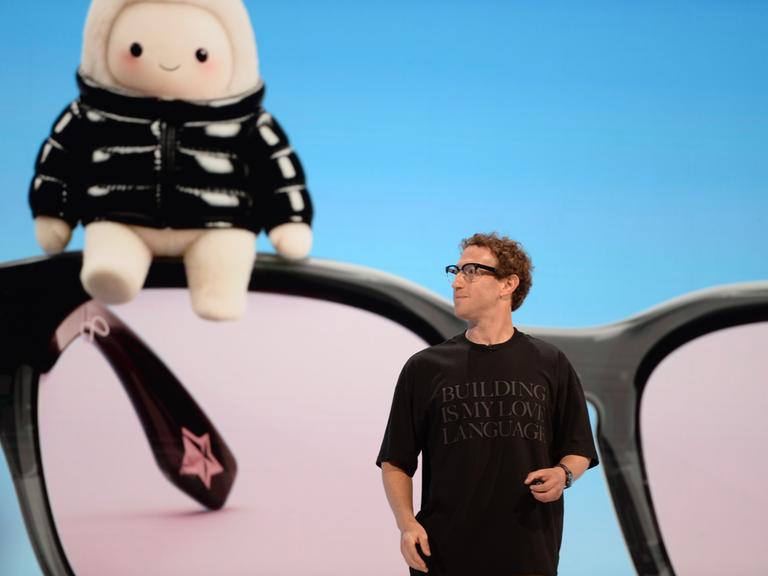 Mark Zuckerberg, Chef des Facebook-Konzerns Meta, steht auf einer Bühne und trägt ein schwarzes T-Shirt mit einer Aufschrift. Hinter ihm ist auf einem Bildschirm ein riesiges Wesen zu sehen, das an ein Tamagotchi oder ein Labubu erinnert.