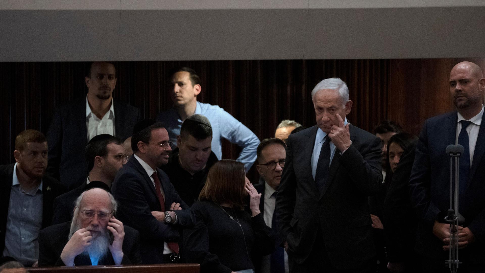 Netanjahu steht im Parlament, um ihn herum stehen weitere Politiker. Netanjahu kratzt sich am Auge.