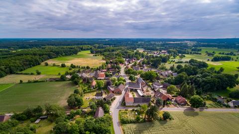 Entlang einer Straße liegen wenige Häuser in einer Landschaft aus Feldern und Wald. 