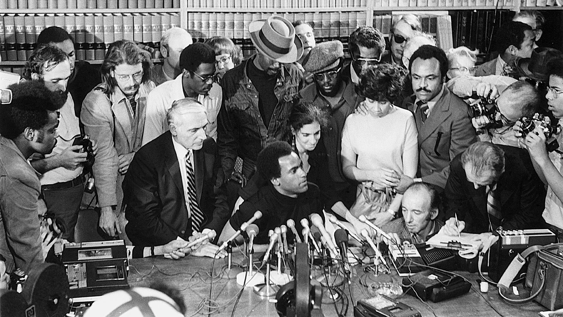 Der US-Aktivist Huey Newton sitzt bei einer Pressekonferenz 1971 an einem Tisch mit einer Reihe von Mikrofonen und wird umringt von einer Gruppe von Menschen