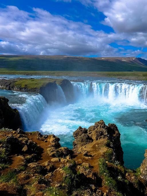 Blick auf einen großen isländischen Wasserfall mit blauem Himmel und wilden Wolken.