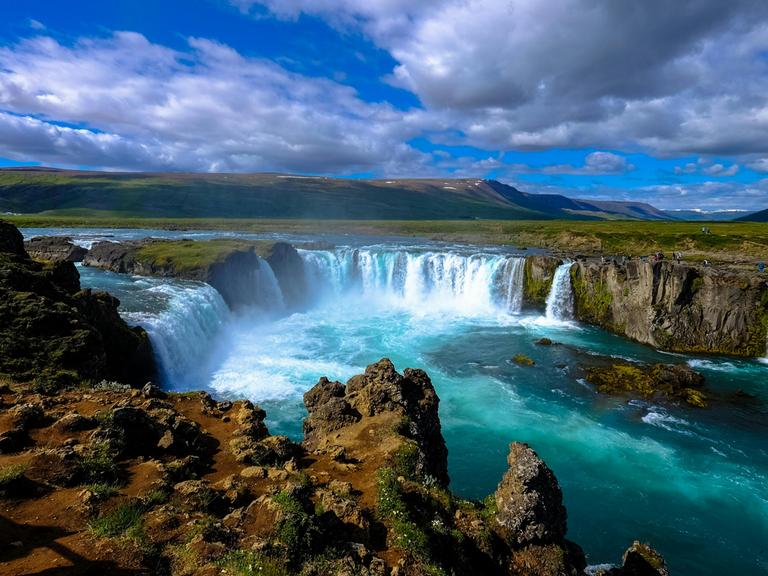 Blick auf einen großen isländischen Wasserfall mit blauem Himmel und wilden Wolken.