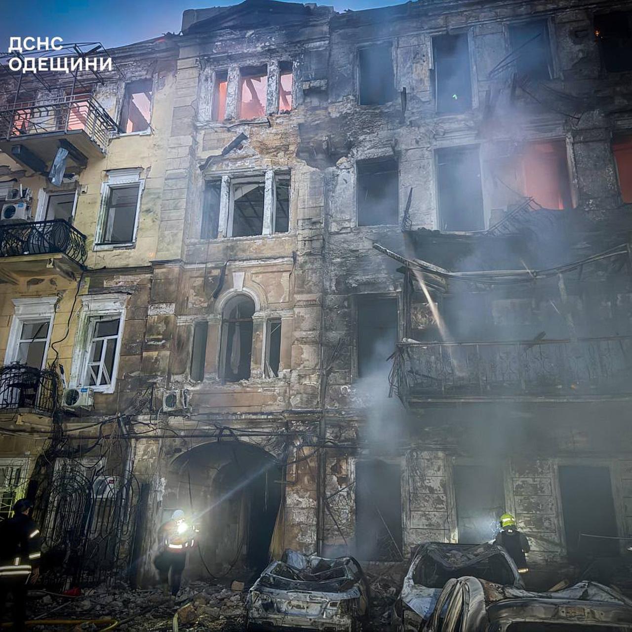 Es ist Nacht. Rettungskräfte stehen vor einem ausgebrannten mehrstöckigen Haus. Davor stehen ausgebrannte Autos. Rauch liegt in der Luft. 