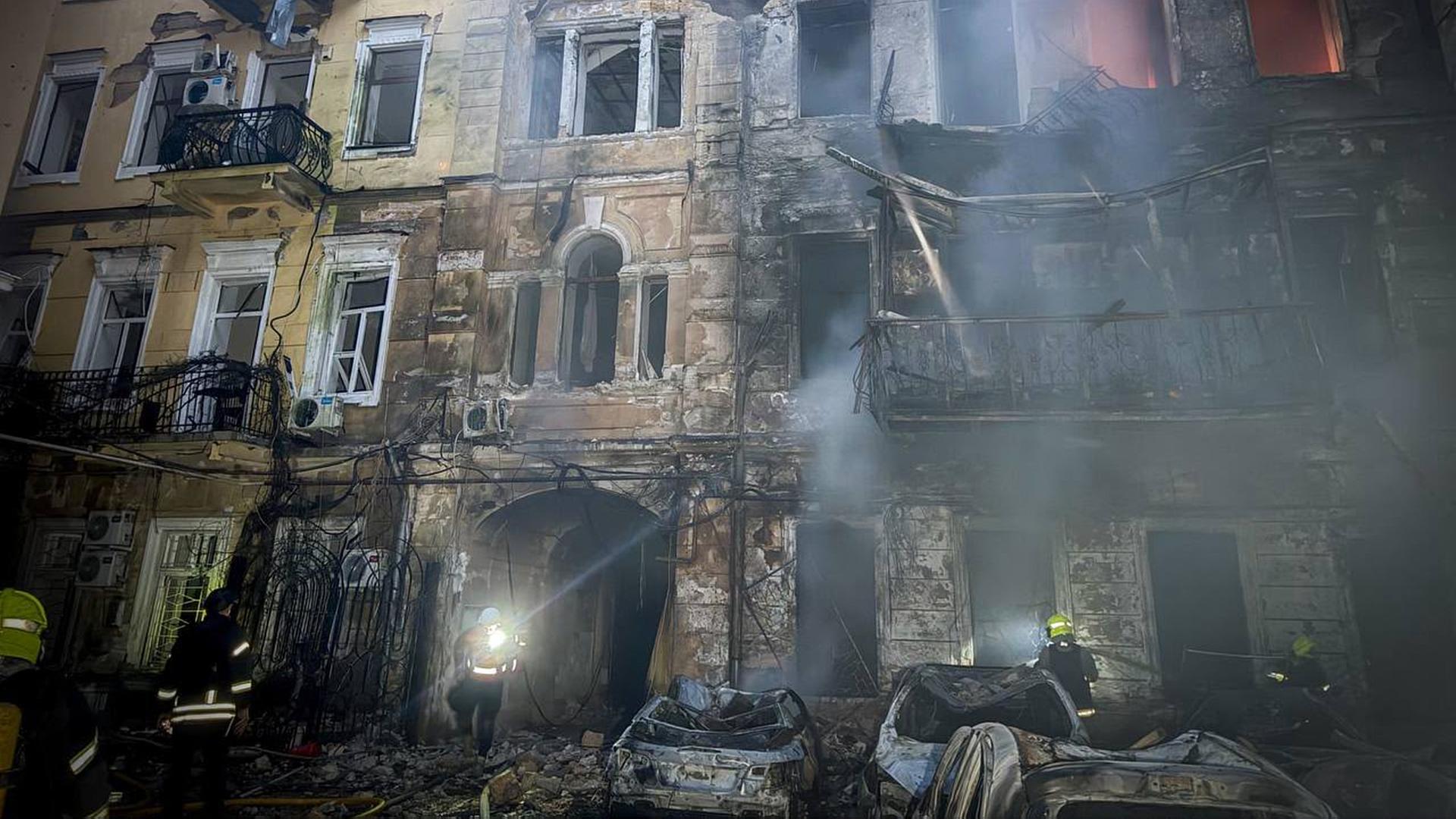 Es ist Nacht. Rettungskräfte stehen vor einem ausgebrannten mehrstöckigen Haus. Davor stehen ausgebrannte Autos. Rauch liegt in der Luft. 