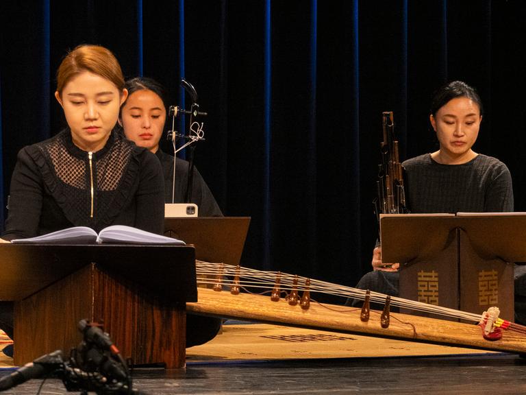 Drei Musikerinnen mit traditionellen koreanischen Instrumenten sitzen mit kontrollierten Blicken auf der Bühne