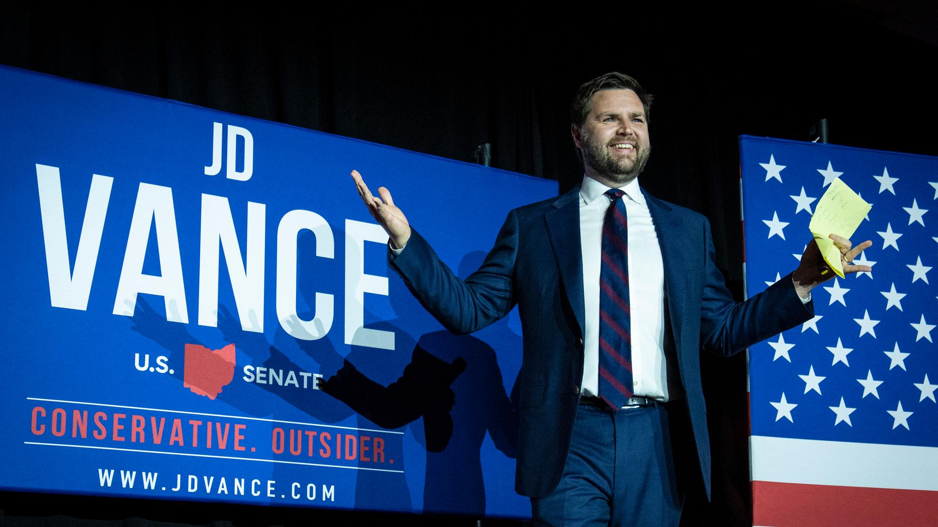 J.D. Vance steht auf einer Bühne. Im Hintergrund erscheint sein Name und ein Ausschnitt der US-Flagge.