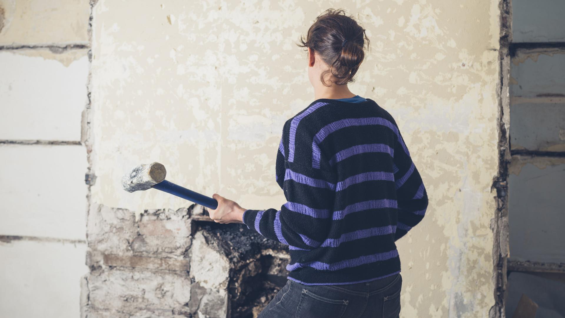 Eine Frau mit Vorschlaghammer ist von hinten zu sehen. Sie steht vor einer alten tapezierten Wand in einem Haus. Eine Frau mit Vorschlaghammer ist von hinten zu sehen. Sie steht vor einer alten tapezierten Wand in einem Haus.