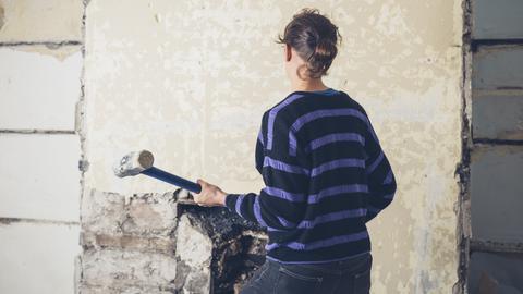 Eine Frau mit Vorschlaghammer ist von hinten zu sehen. Sie steht vor einer alten tapezierten Wand in einem Haus.
