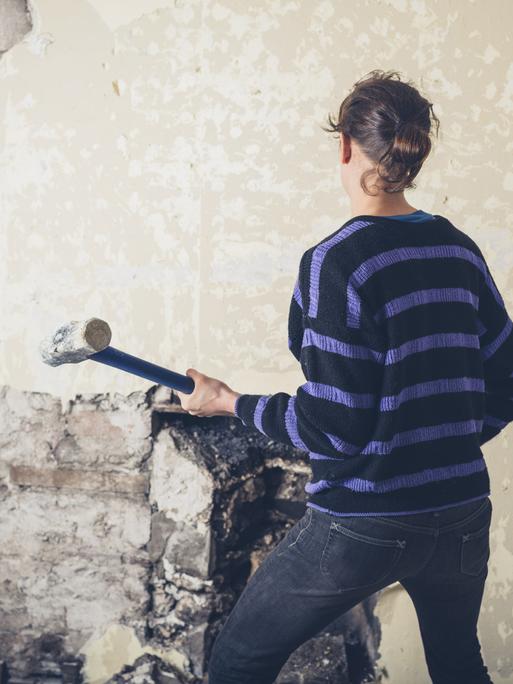 Eine Frau mit Vorschlaghammer ist von hinten zu sehen. Sie steht vor einer alten tapezierten Wand in einem Haus.