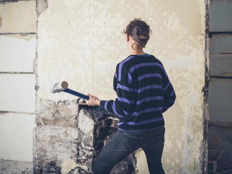Eine Frau mit Vorschlaghammer ist von hinten zu sehen. Sie steht vor einer alten tapezierten Wand in einem Haus.