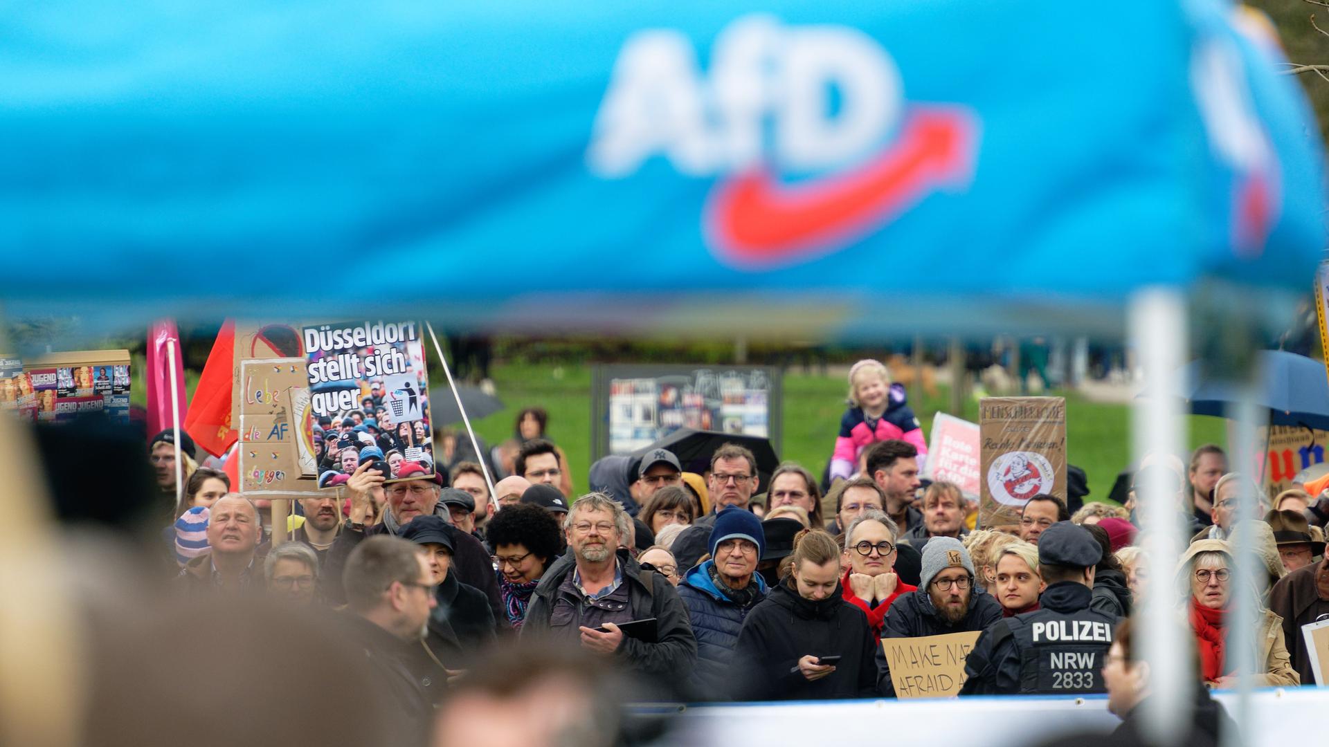 Viele Menschen stehen bei einer Demonstration in Düsseldorf hinter eine Polizeiabsperrung zusammen und halten Plakate hoch. Im Vordergrund ist ein Pavillon mit dem Logo der AfD.