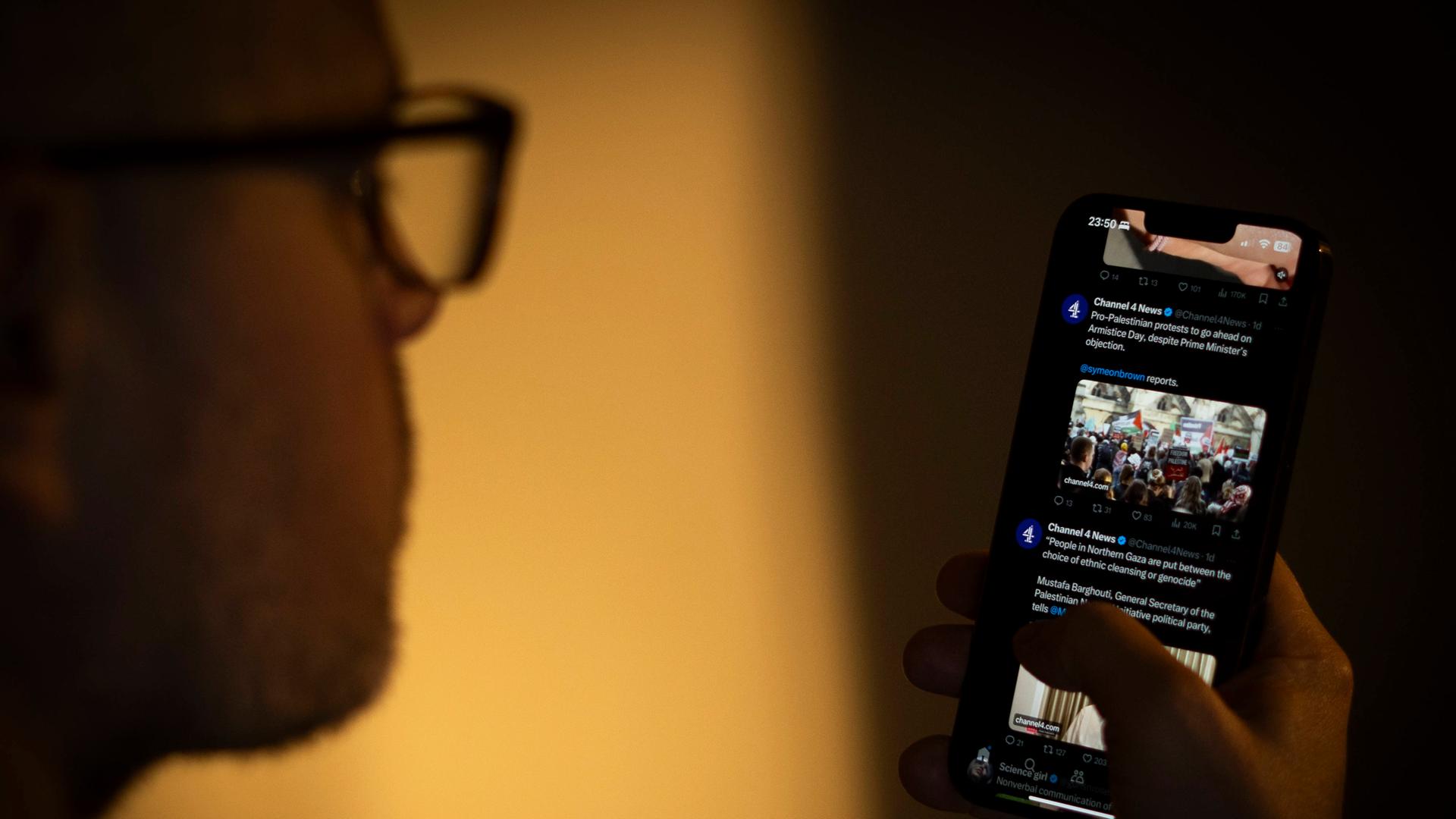 Ein Mensch mit Brille hält ein Smartphone in der Hand und schaut darauf. Auf dem Bildschirm ist die Online-Plattform X geöffnet. 