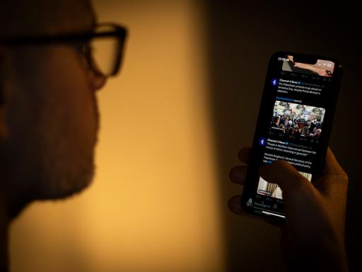 Ein Mensch mit Brille hält ein Smartphone in der Hand und schaut darauf. Auf dem Bildschirm ist die Online-Plattform X geöffnet. 