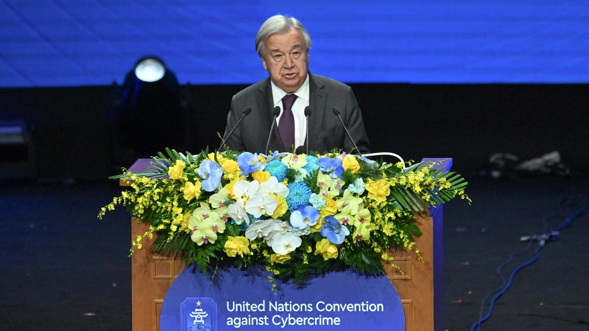 Antonio Guterres steht an einem Rednerpult, das mit Blumen geschmückt ist. Darunter steht "United Nations Convention Against Cybercrime".  Antonio Guterres steht an einem Rednerpult, das mit Blumen geschmückt ist. Darunter steht "United Nations Convention Against Cybercrime".
