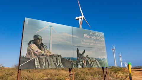 Windräder stehen auf der kolumbianischen Insel La Guajira in einer langen Reihe. Ein großes Plakat verdeckt sie teilweise. Auf dem Plakat ist eine Angehörige der indigenen Bevölkerung mit Packeseln zu sehen.