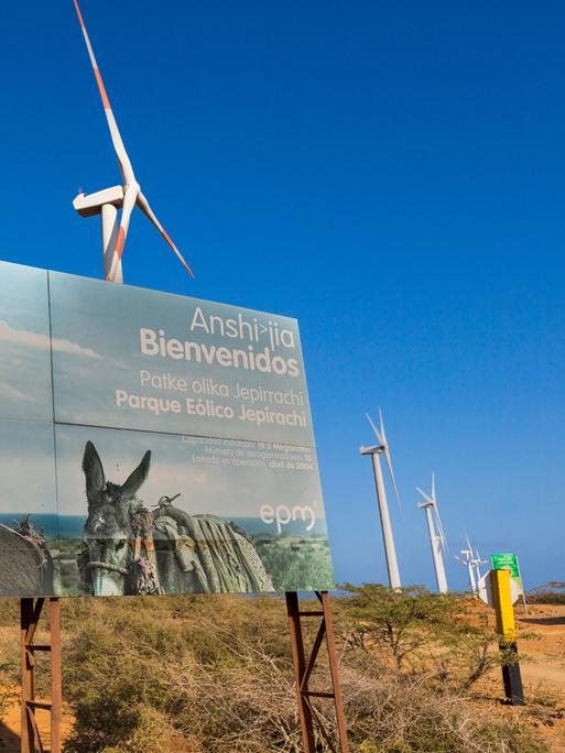 Windräder stehen auf der kolumbianischen Insel La Guajira in einer langen Reihe. Ein großes Plakat verdeckt sie teilweise. Auf dem Plakat ist eine Angehörige der indigenen Bevölkerung mit Packeseln zu sehen.