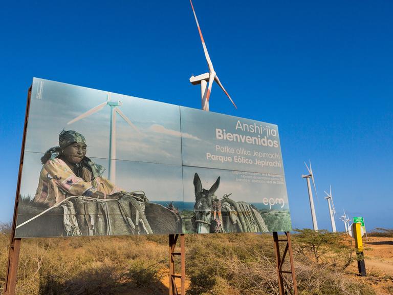 Windräder stehen auf der kolumbianischen Insel La Guajira in einer langen Reihe. Ein großes Plakat verdeckt sie teilweise. Auf dem Plakat ist eine Angehörige der indigenen Bevölkerung mit Packeseln zu sehen.