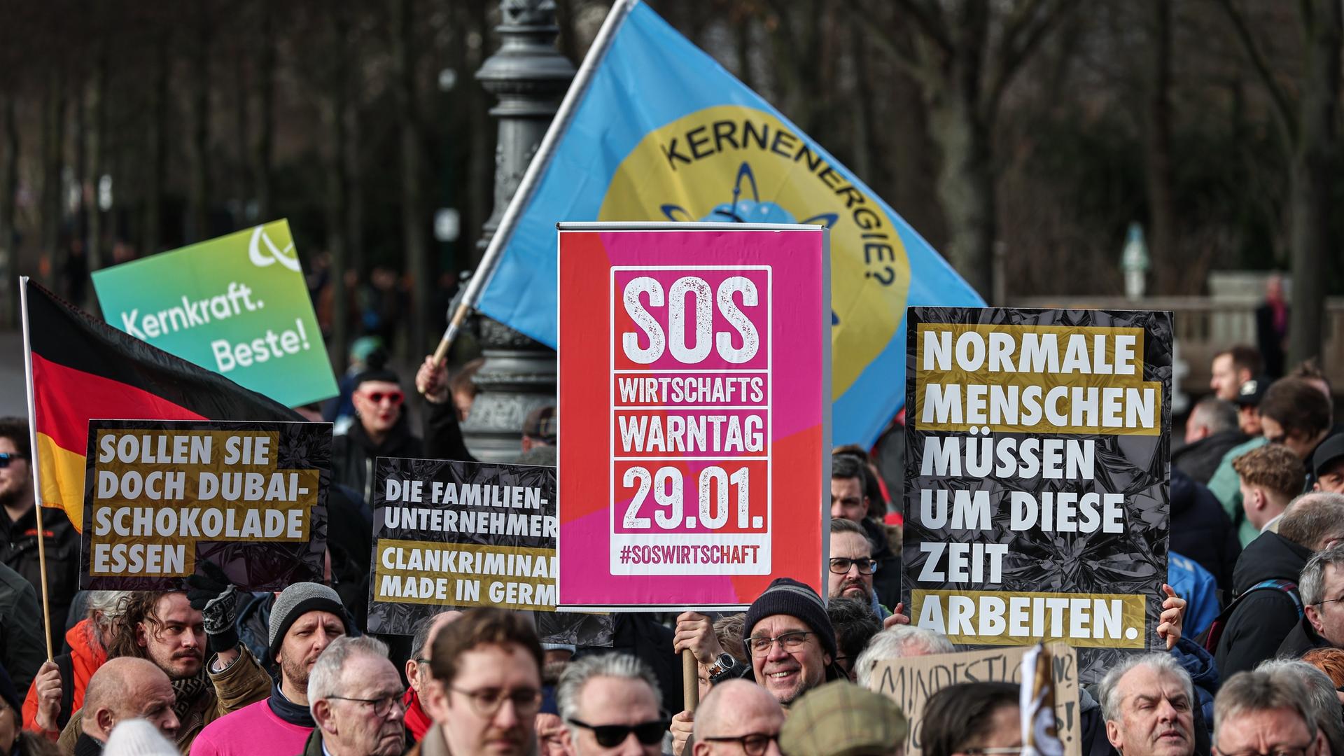 Gegendemonstranten halten dunkle Schilder mit Aufschrift "Normale Menschen müssen um diese Zeit arbeiten" und "Sollen sie doch Dubai-Schokolade essen" auf der Kundgebung "Wirtschaftswarntag" vor dem Brandenburger Tor hoch.