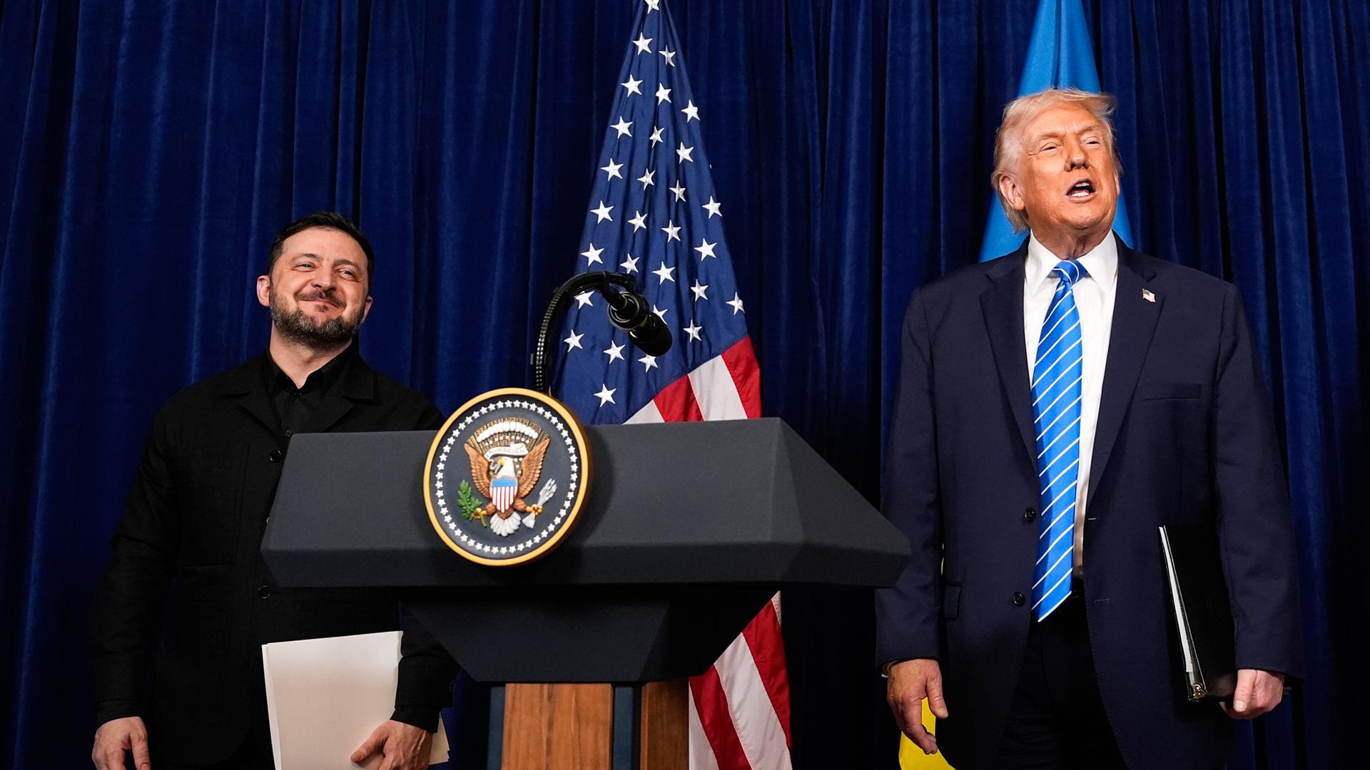Präsident Trump und Präsident Selenskyj stehen auf einem Podium vor einer US-Flagge. Trump redet, Selenskyj lächelt.