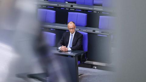 Bundeskanzler Friedrich Merz sitzt allein mit einem ernsten Gesicht auf einem blauen Stuhl im Bundestag.