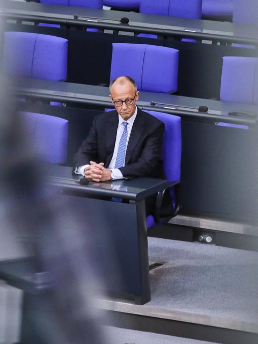 Bundeskanzler Friedrich Merz sitzt allein mit einem ernsten Gesicht auf einem blauen Stuhl im Bundestag.