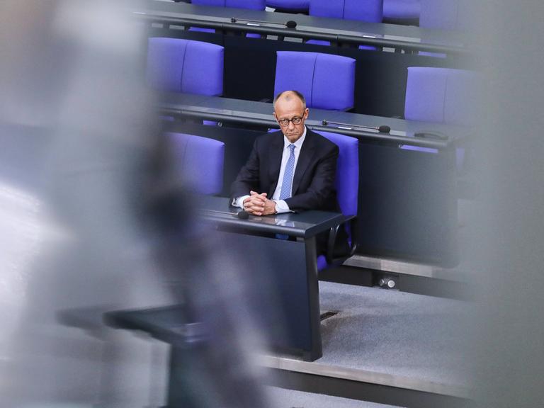 Bundeskanzler Friedrich Merz sitzt allein mit einem ernsten Gesicht auf einem blauen Stuhl im Bundestag.