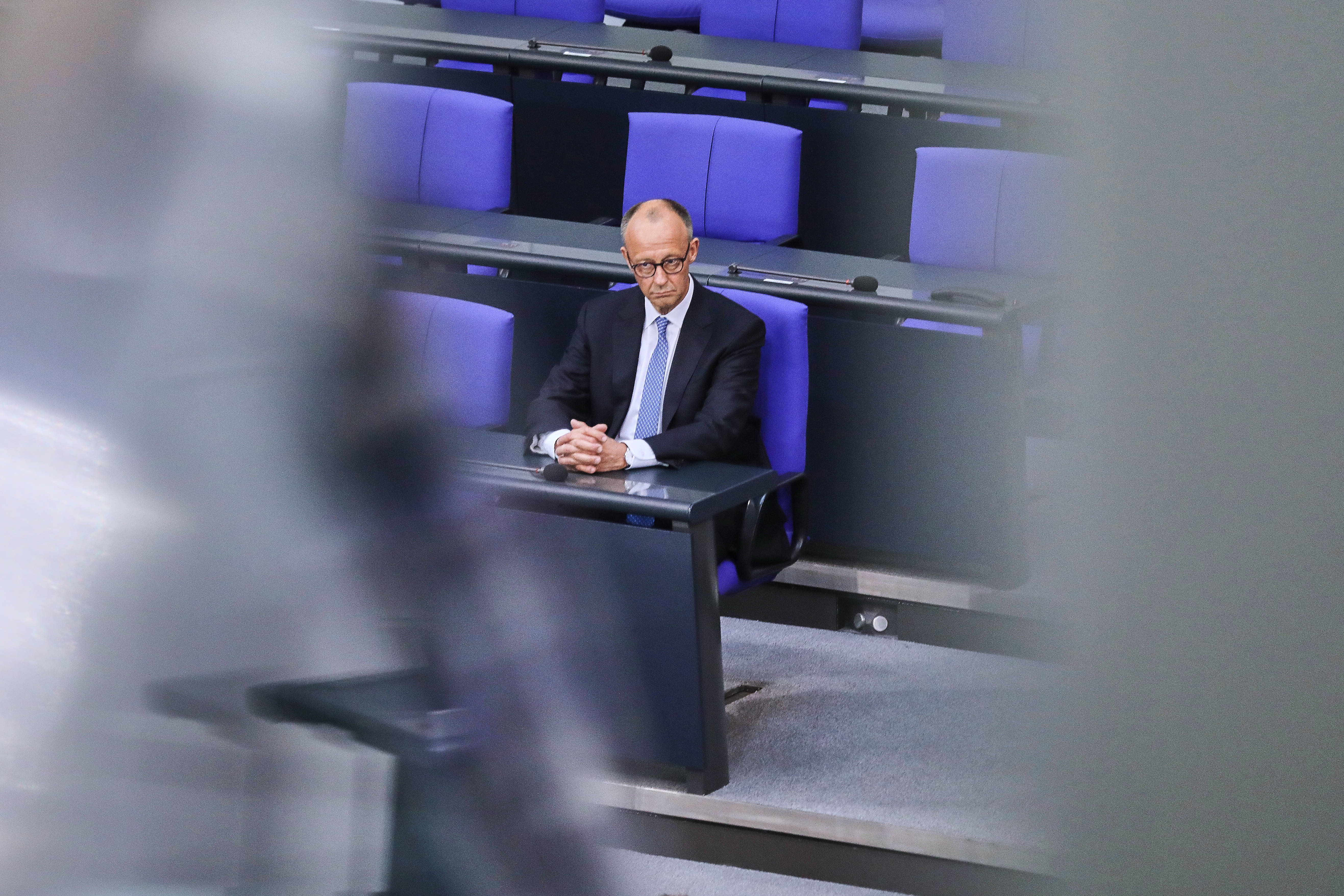 Bundeskanzler Friedrich Merz sitzt allein mit einem ernsten Gesicht auf einem blauen Stuhl im Bundestag.