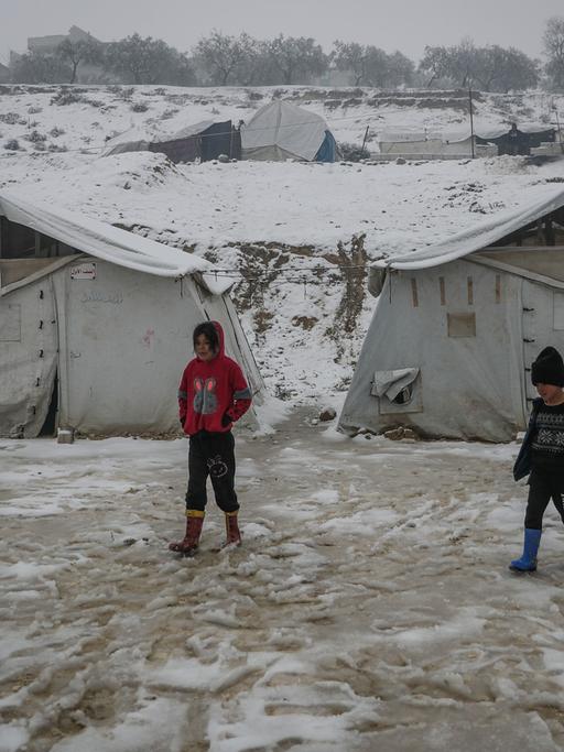 Drei Kinder stehen vor Zelten in einem Flüchtlingslager. Es liegt Schnee.