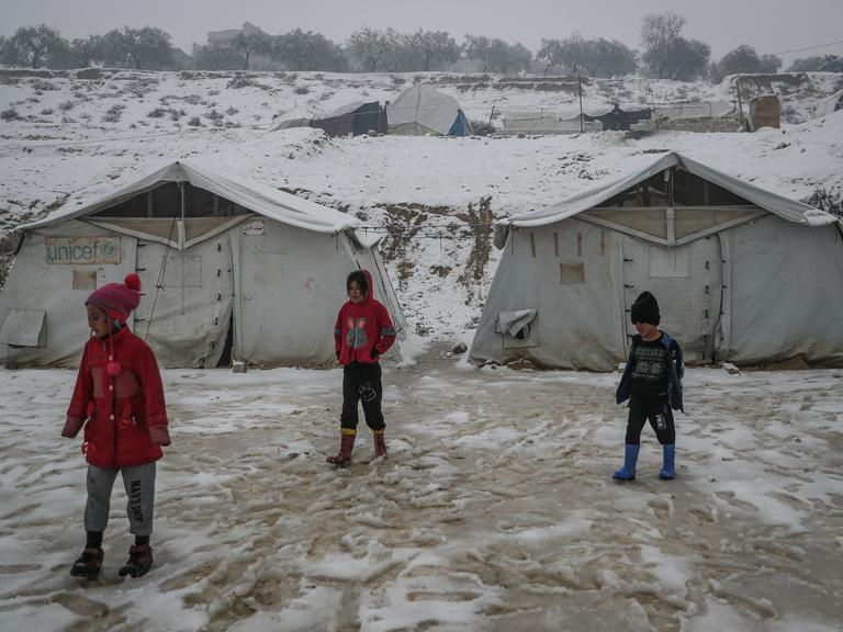 Drei Kinder stehen vor Zelten in einem Flüchtlingslager. Es liegt Schnee.