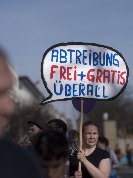 Eine Frau trägt ein Schild mit der Aufschrift: "Abtreibung frei + gratis überall". Im Vordergrund des Bildes ist unscharf ein Mann zu sehen, der den Kopf so hält, als schaue er direkt auf das Schild.