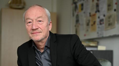 Helmut Eignersitzt im Büro in lässiger Pose am Schreibtisch. Er trägt ein schwarzes Sakko über grauem Hemd.
