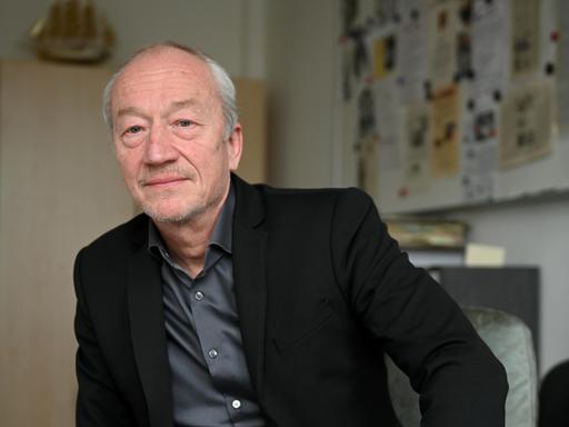 Helmut Eignersitzt im Büro in lässiger Pose am Schreibtisch. Er trägt ein schwarzes Sakko über grauem Hemd.