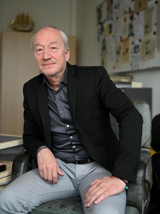 Helmut Eignersitzt im Büro in lässiger Pose am Schreibtisch. Er trägt ein schwarzes Sakko über grauem Hemd.