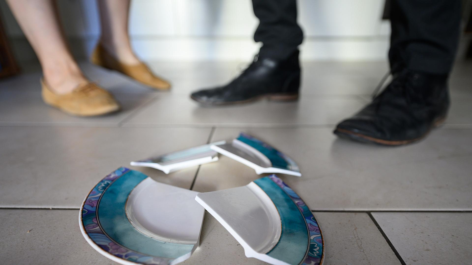 Füße eines Mannes und einer Frau hinter einem zerbrochenen Teller in einer Küche.