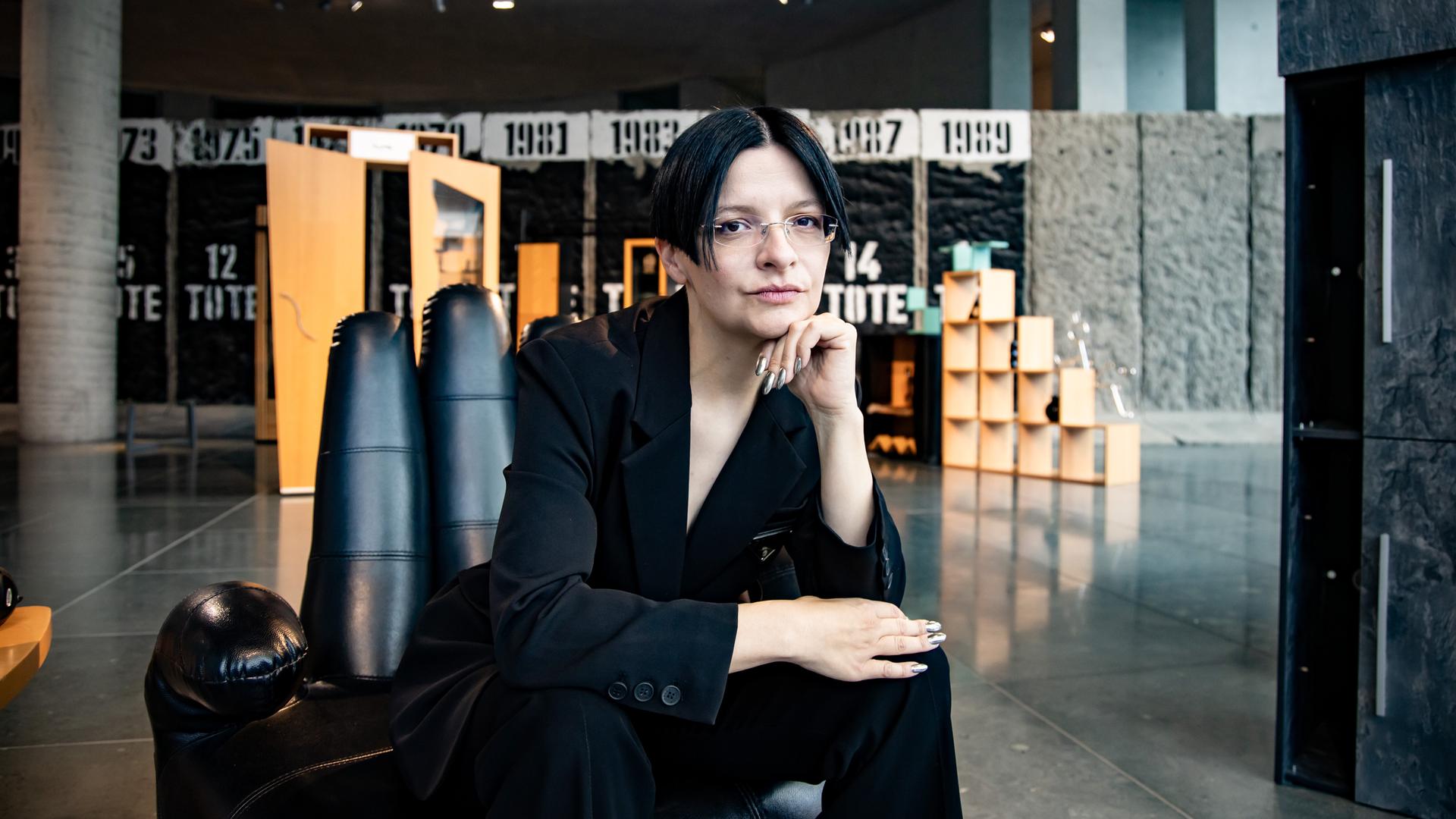 Eine Frau mit dunklen Haaren sitzt in einem schwarzen Anzug auf einem Sessel in Form einer Hand. 