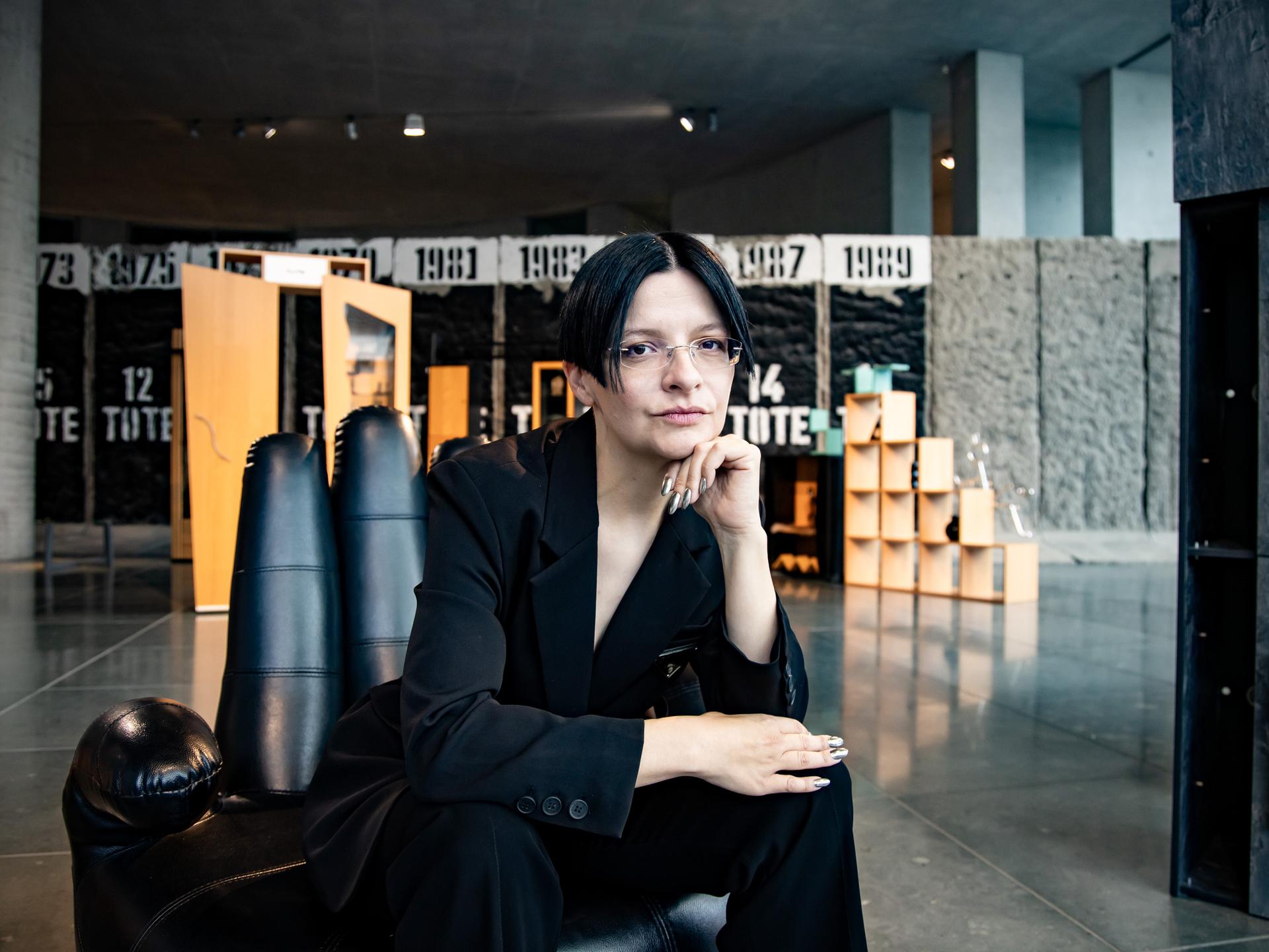 Eine Frau mit dunklen Haaren sitzt in einem schwarzen Anzug auf einem Sessel in Form einer Hand. 