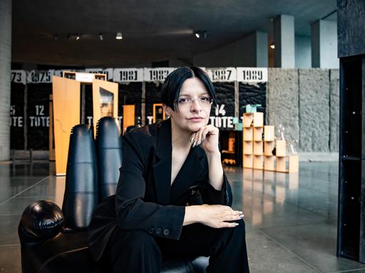 Eine Frau mit dunklen Haaren sitzt in einem schwarzen Anzug auf einem Sessel in Form einer Hand. 