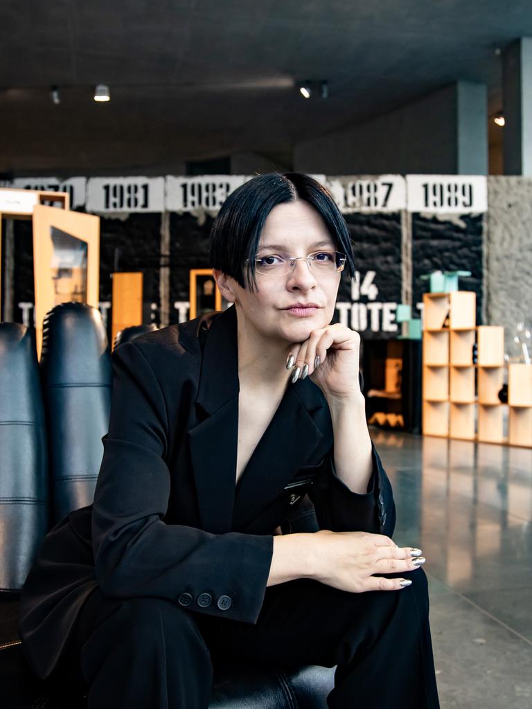 Eine Frau mit dunklen Haaren sitzt in einem schwarzen Anzug auf einem Sessel in Form einer Hand. 