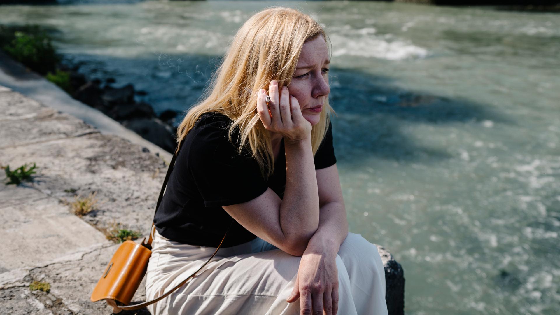 Sandra Hüller als Ingeborg Bachmann sitzt, nachdenklich auf das Wasser schauend und rauchend, am Ufer des Tiber in Rom.