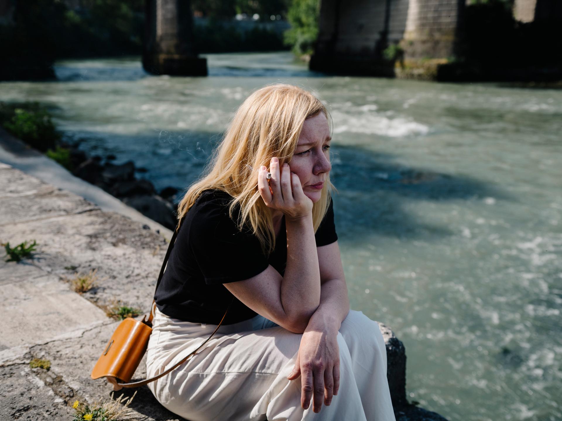 Sandra Hüller als Ingeborg Bachmann sitzt, nachdenklich auf das Wasser schauend und rauchend, am Ufer des Tiber in Rom.