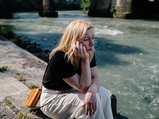 Sandra Hüller als Ingeborg Bachmann sitzt, nachdenklich auf das Wasser schauend und rauchend, am Ufer des Tiber in Rom.