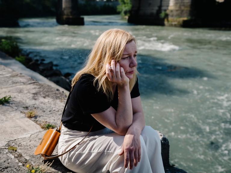 Sandra Hüller als Ingeborg Bachmann sitzt, nachdenklich auf das Wasser schauend und rauchend, am Ufer des Tiber in Rom.
